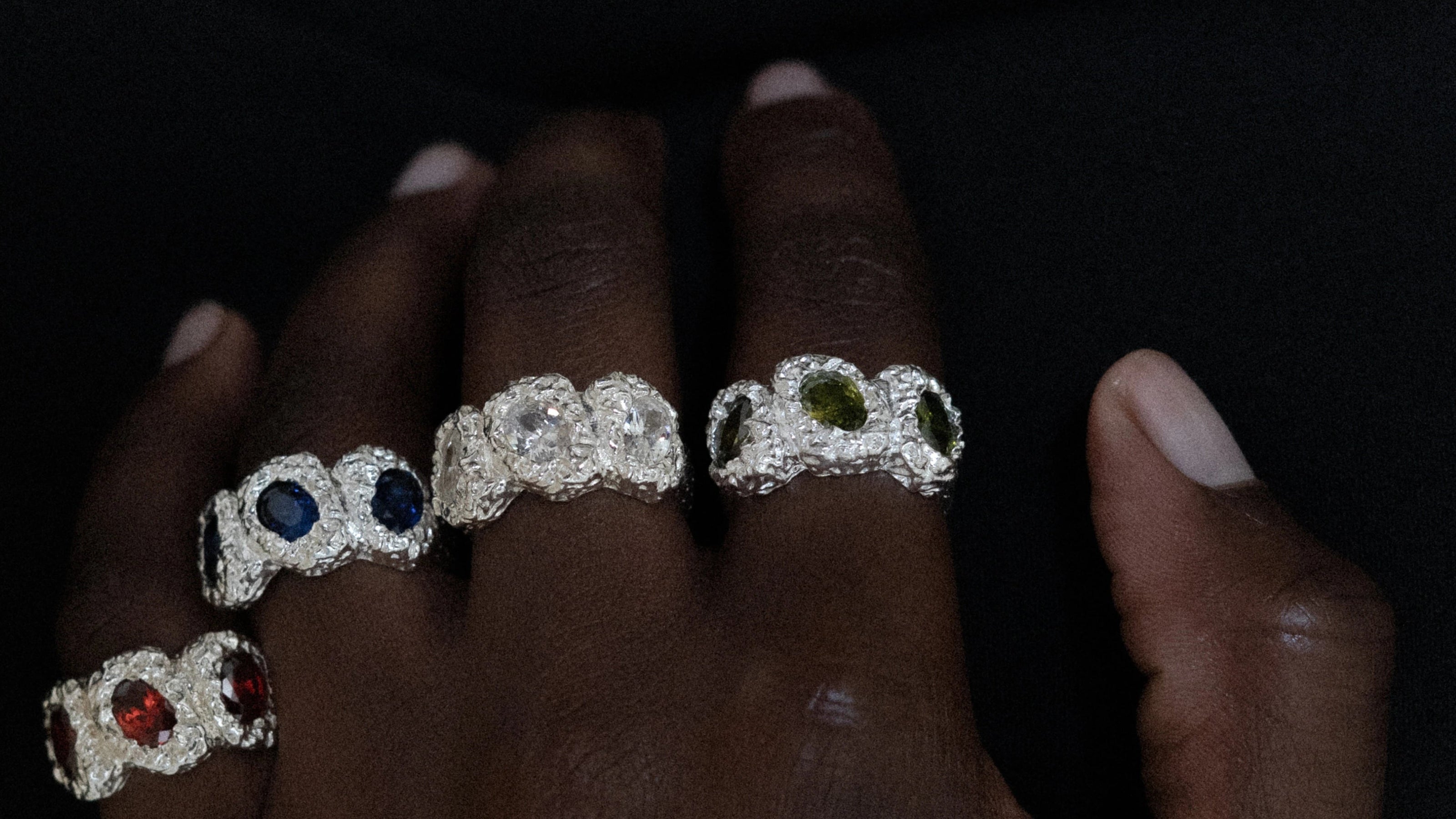 Close-up of a hand wearing multiple diamond rings with colored gemstones on a dark background.