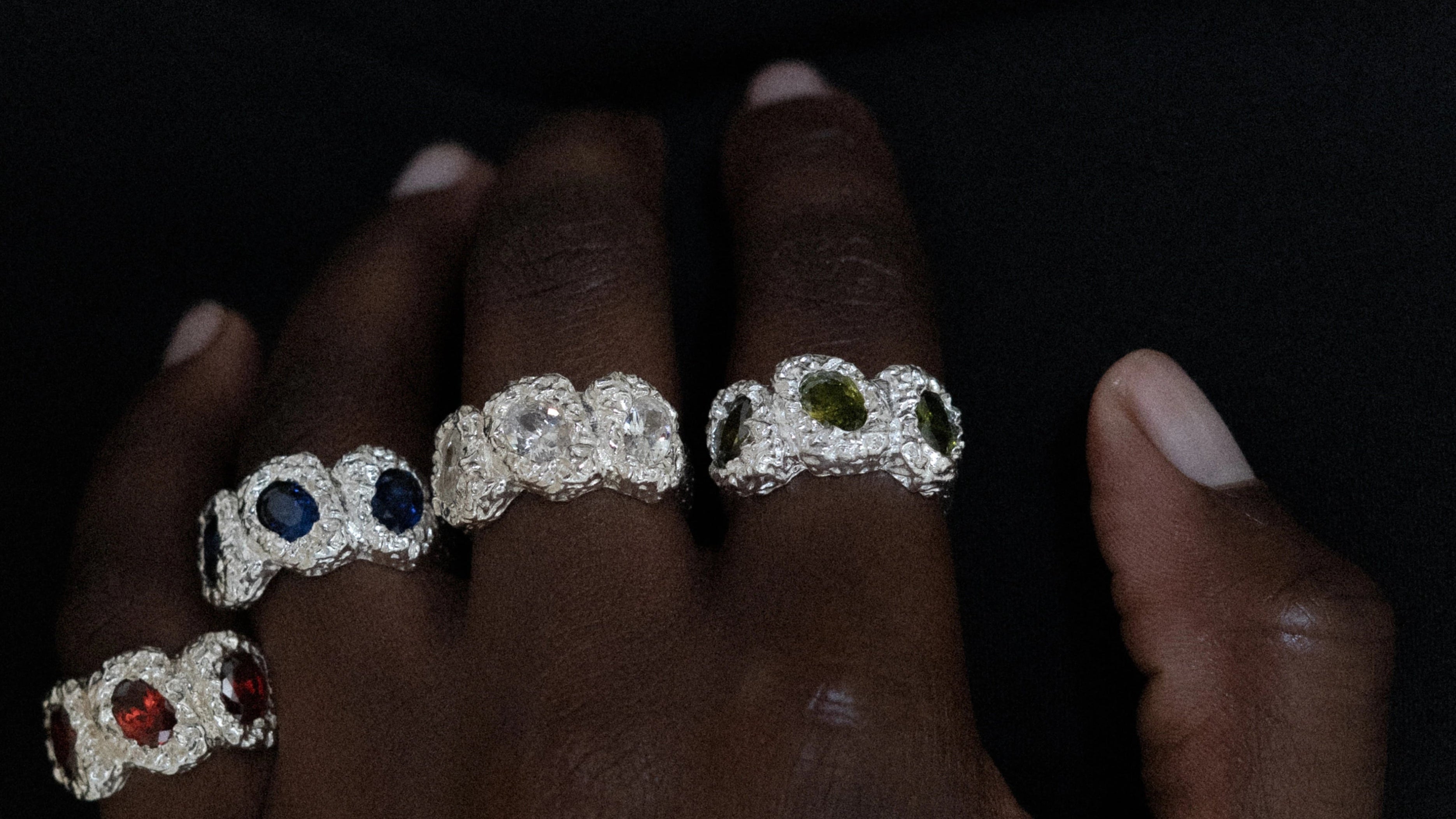 Close-up of a hand wearing multiple diamond rings with colored gemstones on a dark background.slider_item_mBxiQD